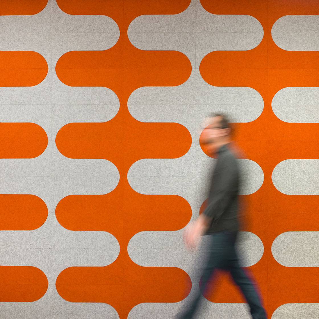 A man blurred by motion walks in front of a felt wall in gray and orange with a curvy 60s style pattern.