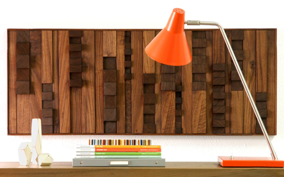 Rectangular wood panel with varying heights is hung on a wall behind a desk with magazines and an orange lamp.