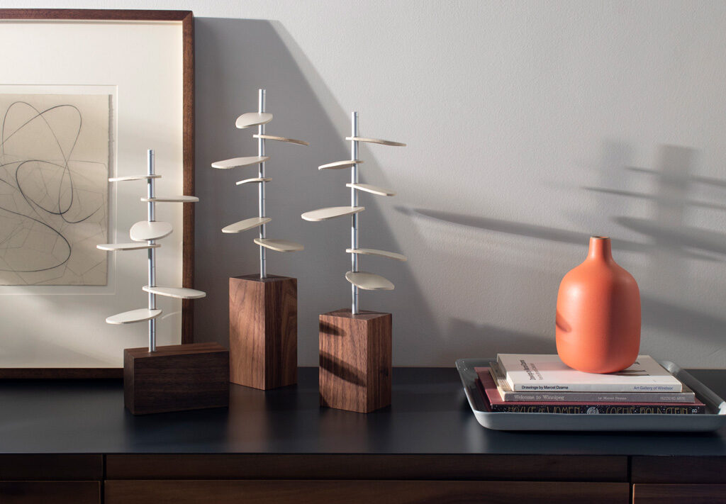 Three tree shaped design pieces made with wood bases, metal stems and white leather leaves sit on a table with books and art