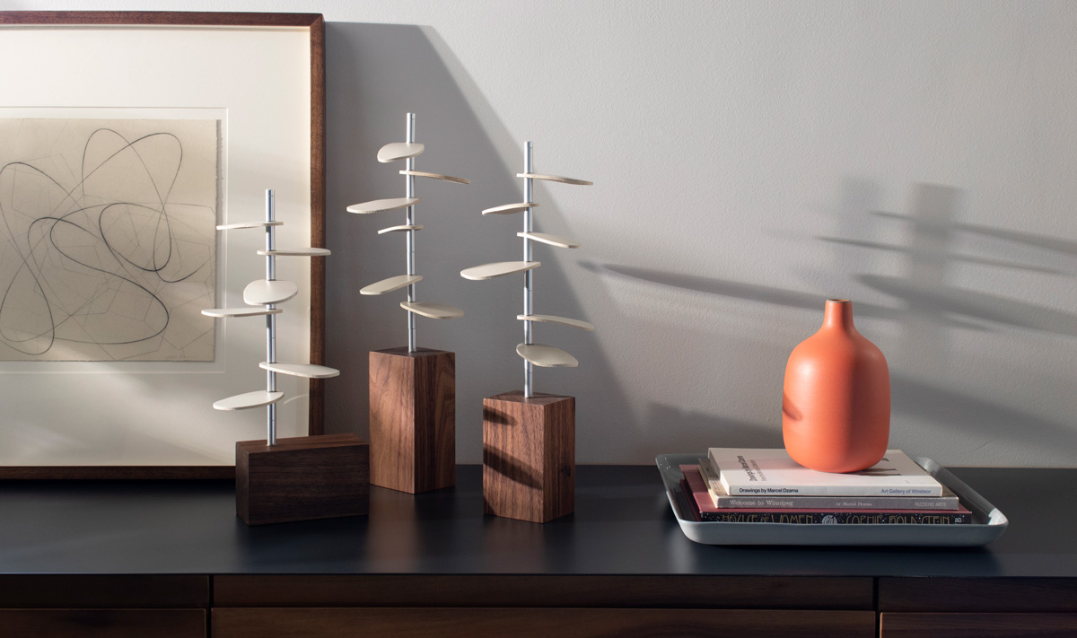 Three tree shaped design pieces made with wood bases, metal stems and white leather leaves sit on a table with books and art