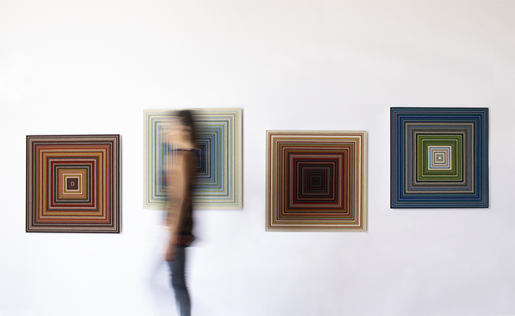 A figure blurred by movement walks in front of 4 square wall panels with concentric square patterns.