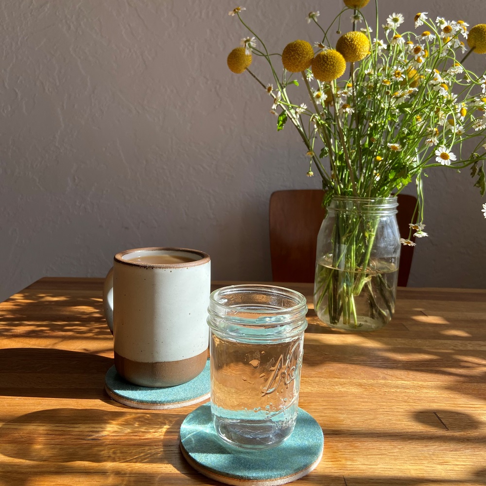 Mug, glass with water both on turquoise felt coasters on a table with daisies in a jar.