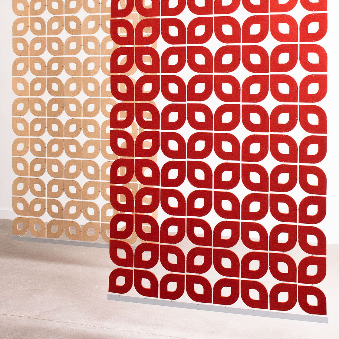 One tan and one red felt screen in an open petal shaped pattern hang in a white space.