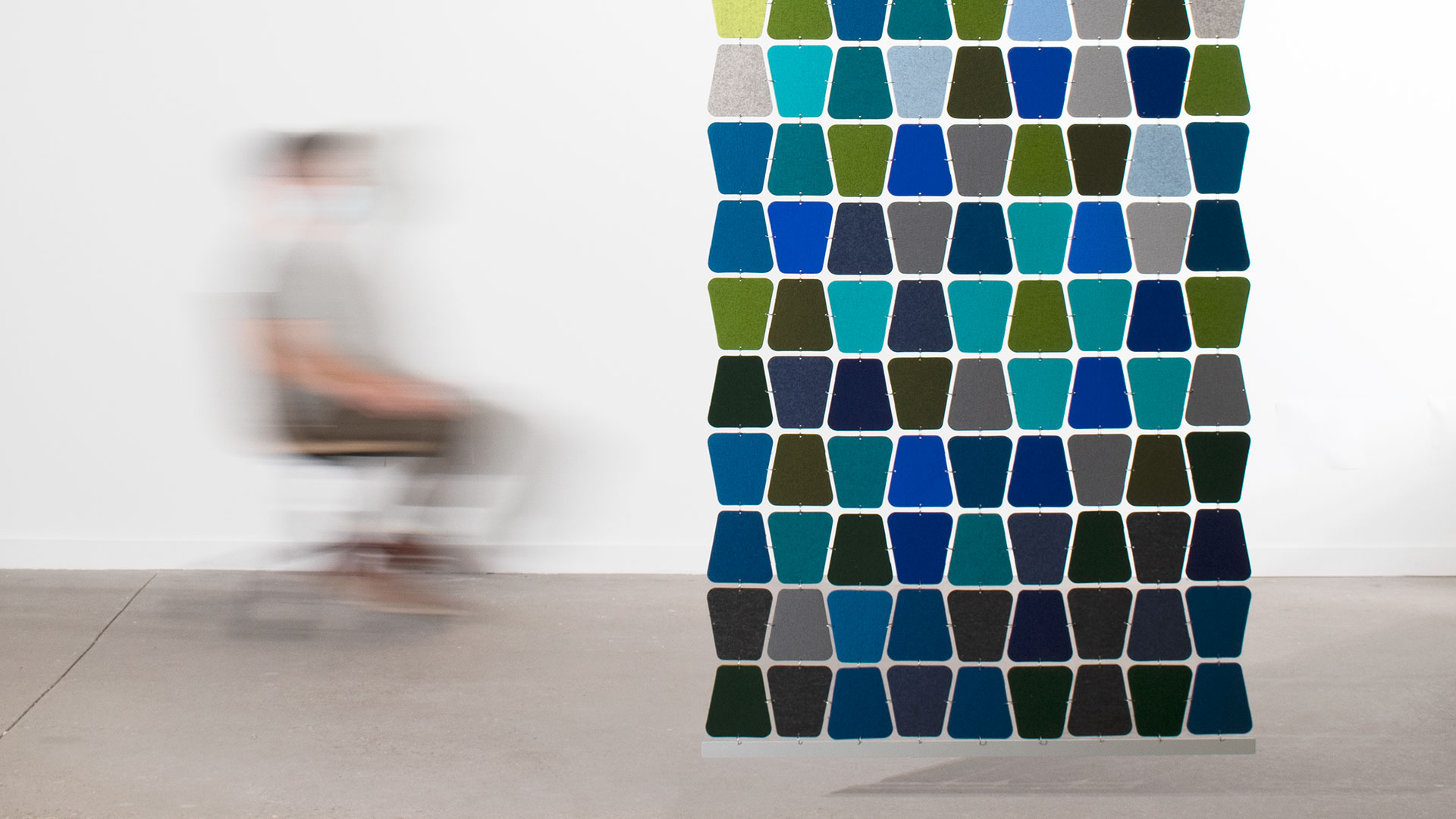 Softly curved trapezoid shaped felt pieces in blues and greens connected with metal rings hangs in a space with a blurry figure in an office chair.