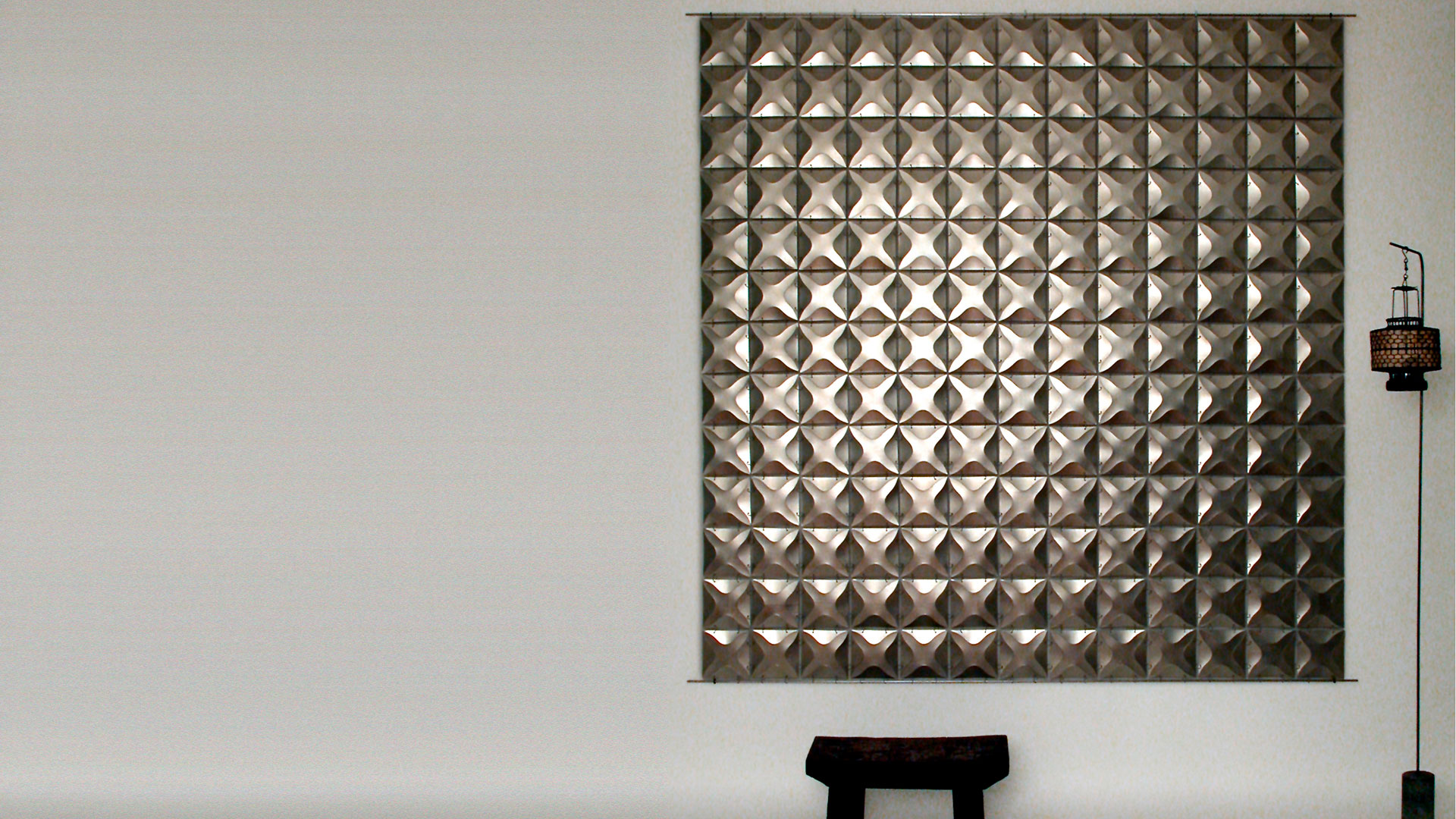 Shiny metal wall hanging constructed of many smaller 3-dimensional x-patten squares next to bench and lamp
