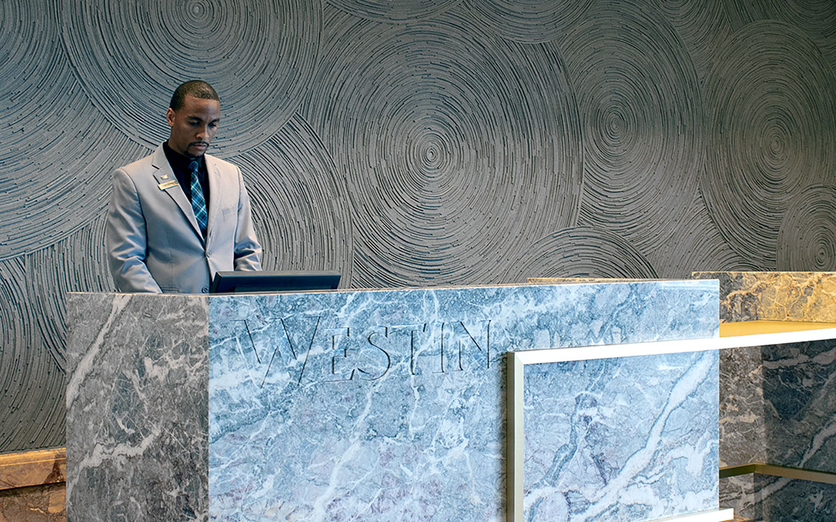 A Black man stands at a gray marble front desk with the word Westin carved into it. Behind him is a gray felt wall with overlapping circular patterns