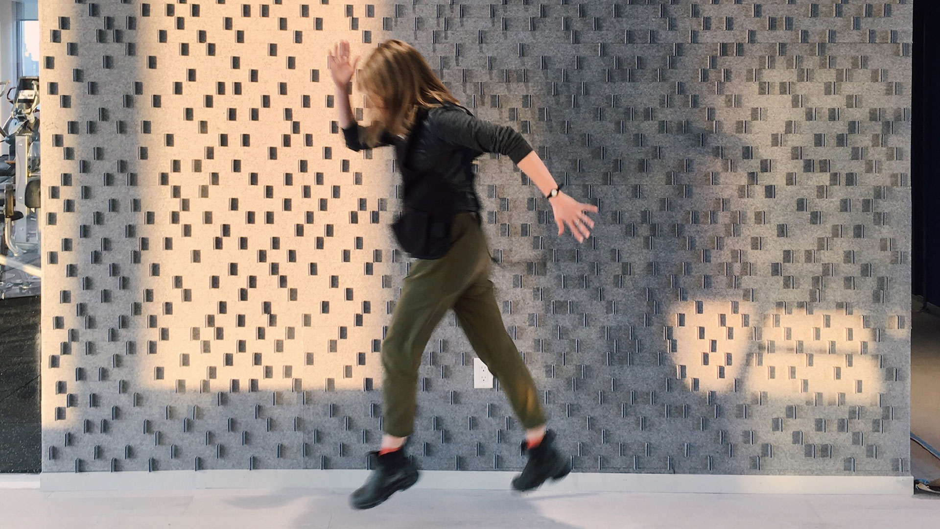 A figure with brown hair, black shirt, and green pants jumps in front of a gray felt wall with shirt vertical felt tabs