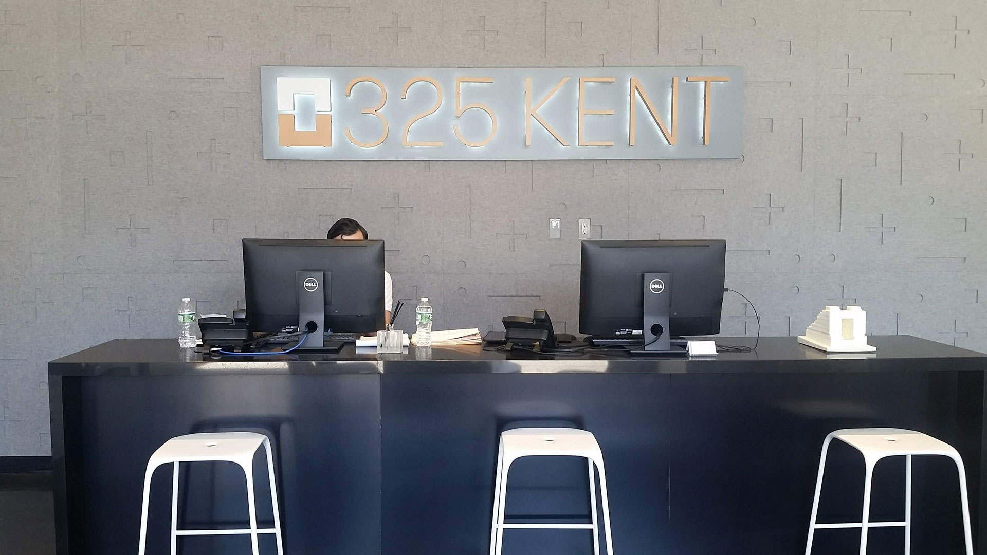 A desk with 3 stools and two computers. The wall behind it has a gray felt wall covering with plus and minus signs and a illuminated sign that says 325 Kent. This is a custom wall covering for a condo building in Brooklyn