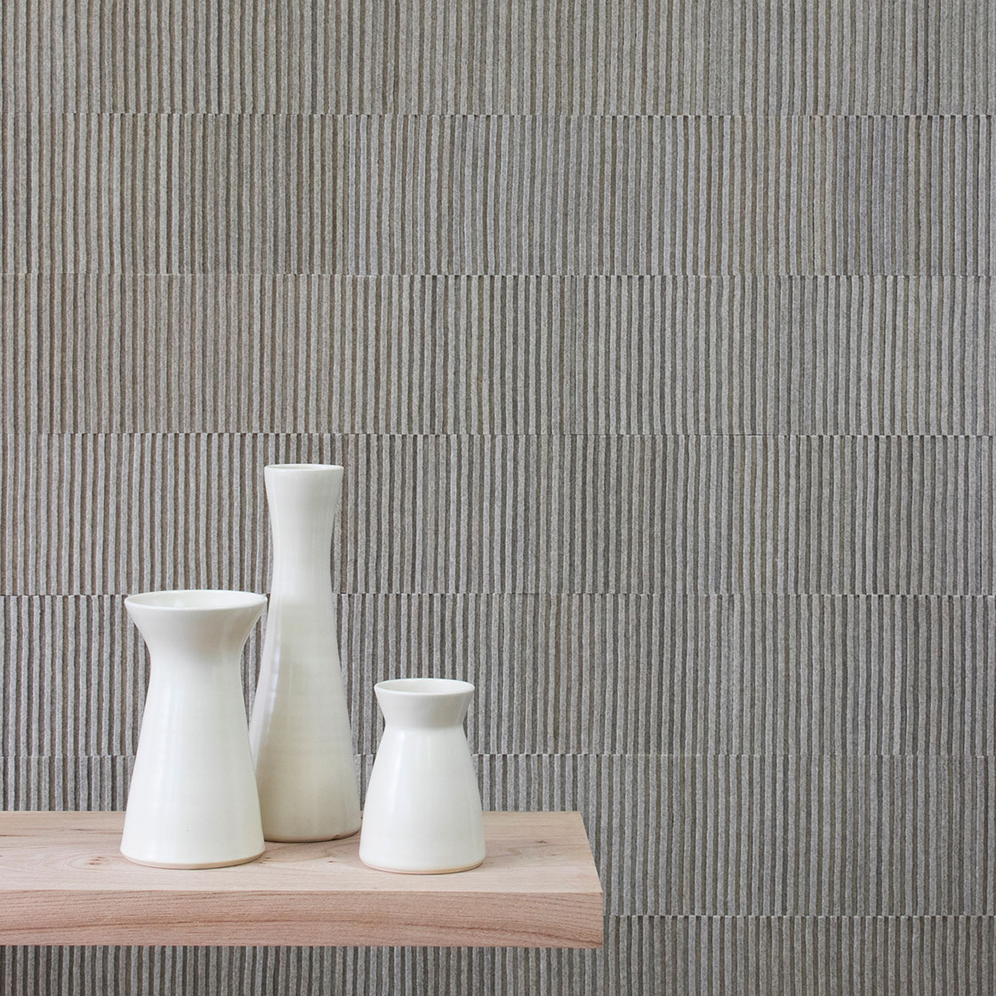Ribbed wall covering in a gray heathered felt. In the foreground are three white ceramic vases on a wood shelf.