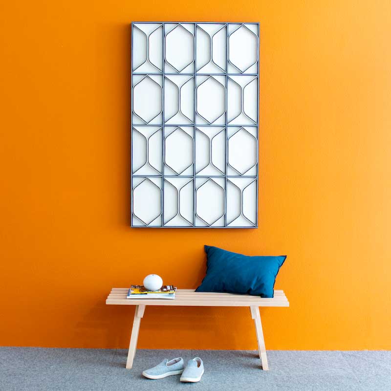 An orange wall with a bench and blue pillow. Above the bench hangs a felt wall art panel with straight lines and subtle diamond shapes. It has a white background and blue lines
