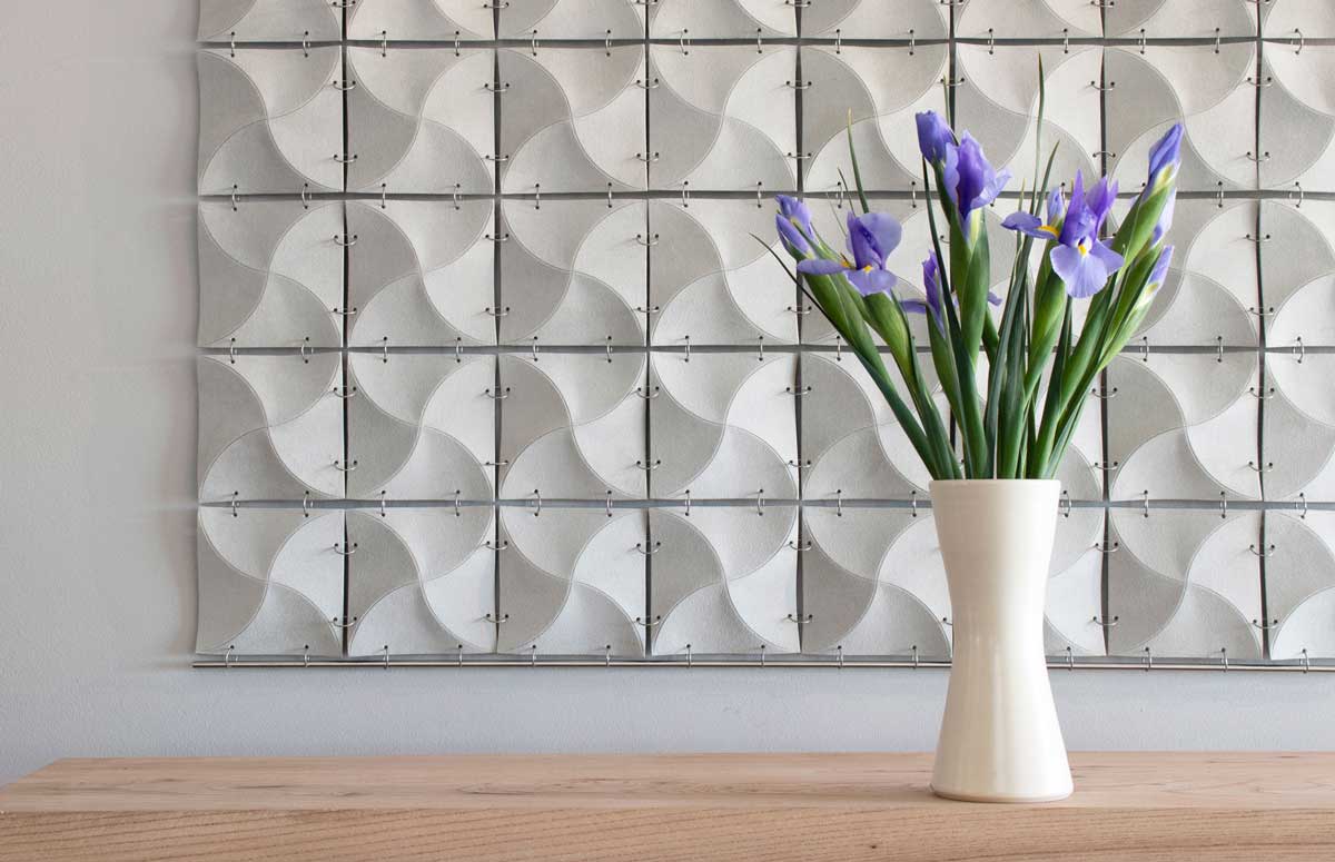 White suede square wall hanging constructed of many smaller wavy squares connected with metal rings hanging behind a white vase with purple irises