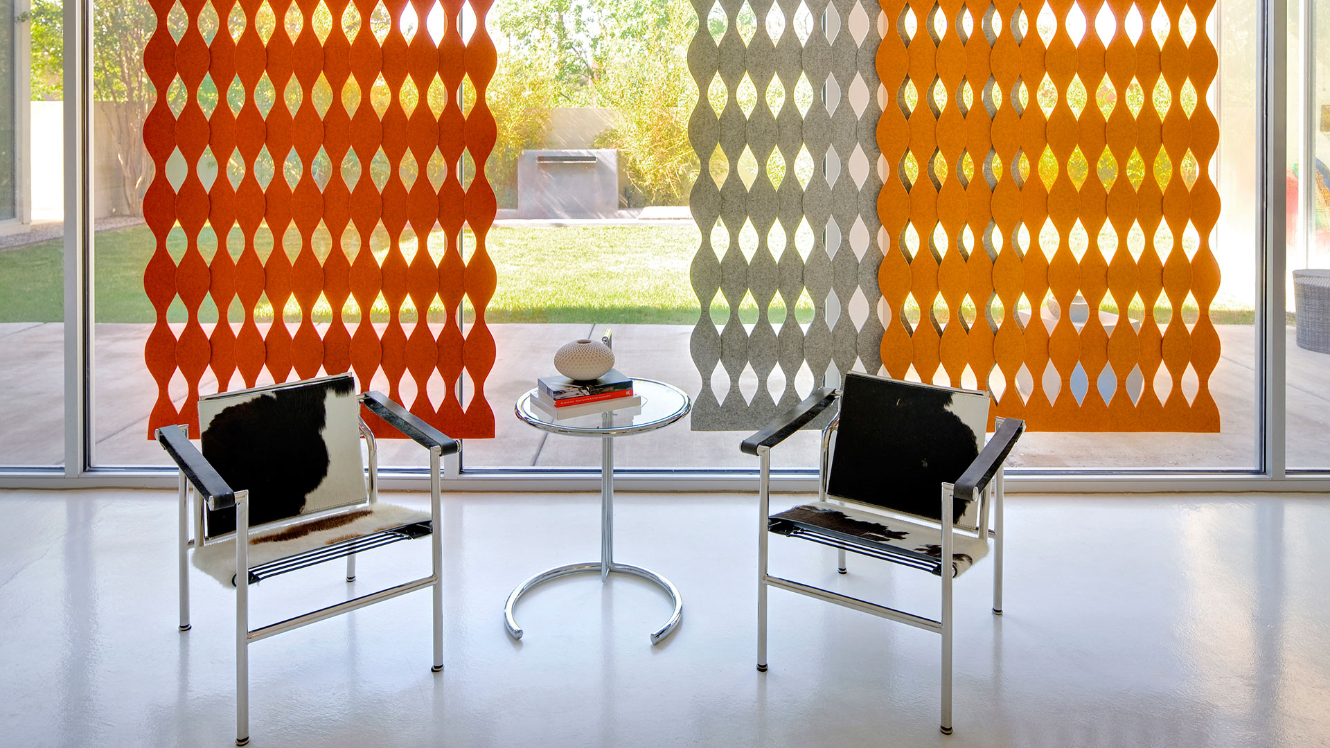 Gray red and orange curvy felt screens next to large window with two cow hide chairs