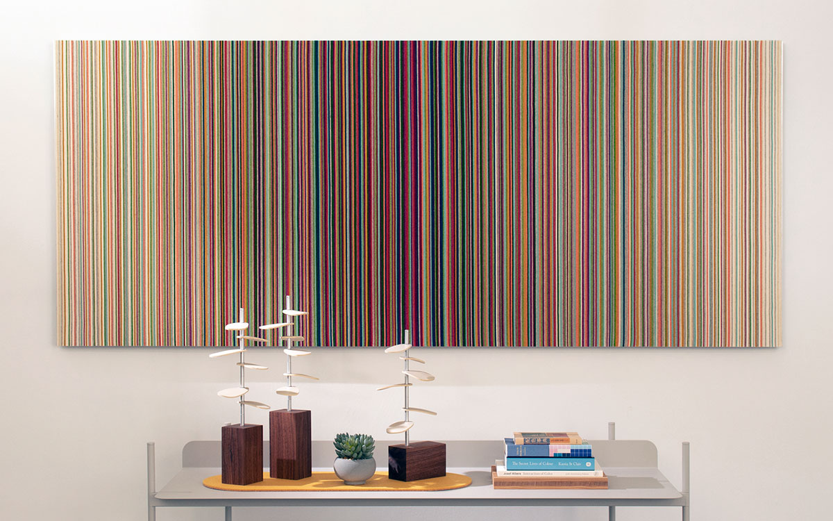 Rectangular felt wall panel with vertical stripes in pink, peach, and blue colors hanging behind a table with books and knick knacks.
