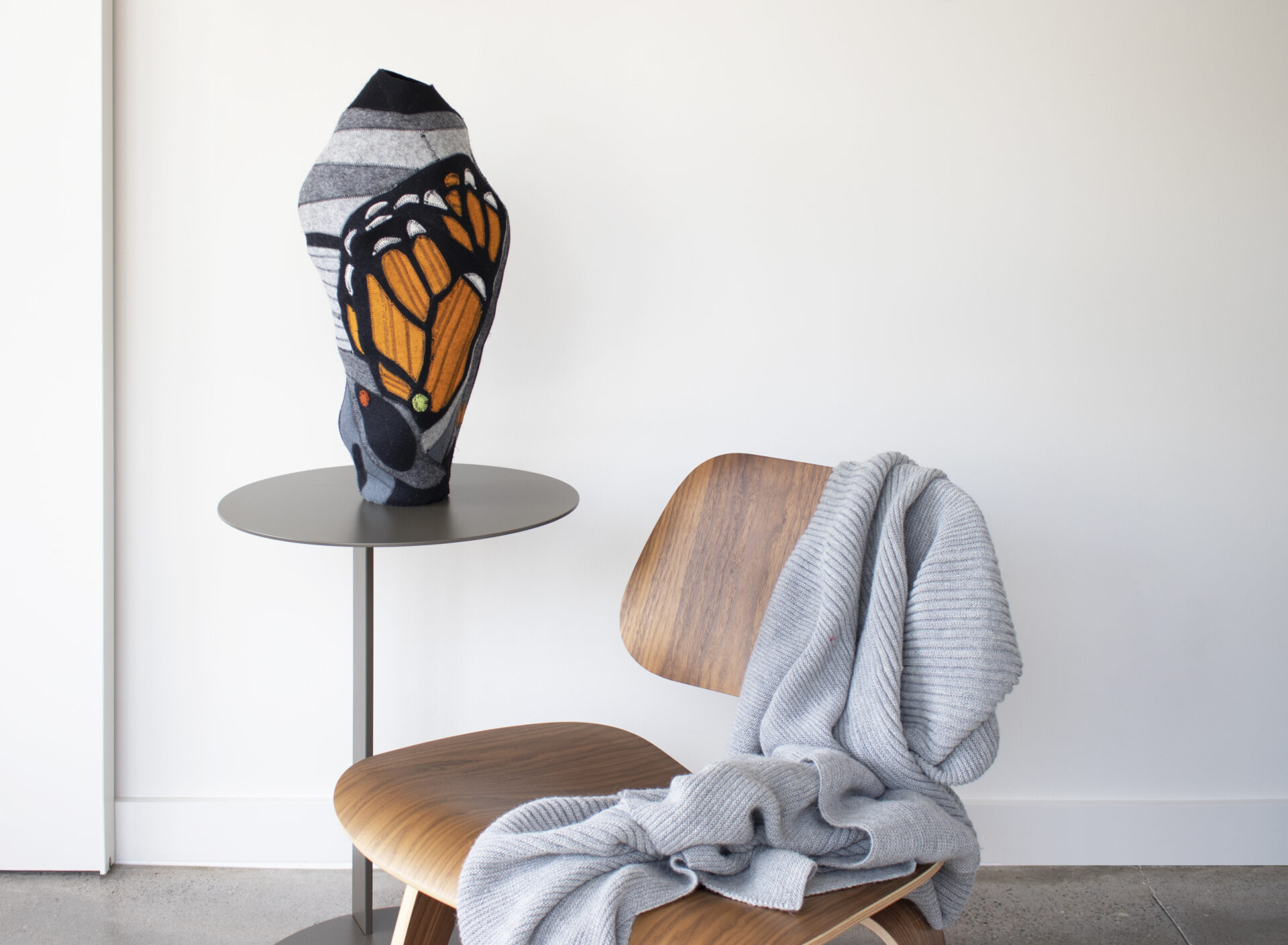 a colorful vase made of wool felt featuring the pattern of a monarch butterfly wing sits on a table beside a bent plywood chair