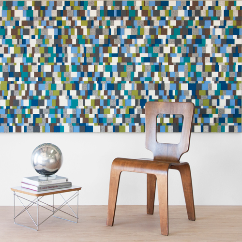 Behind a chair is a large multicolored wall art piece made from small squares of blue, green, and gray felt