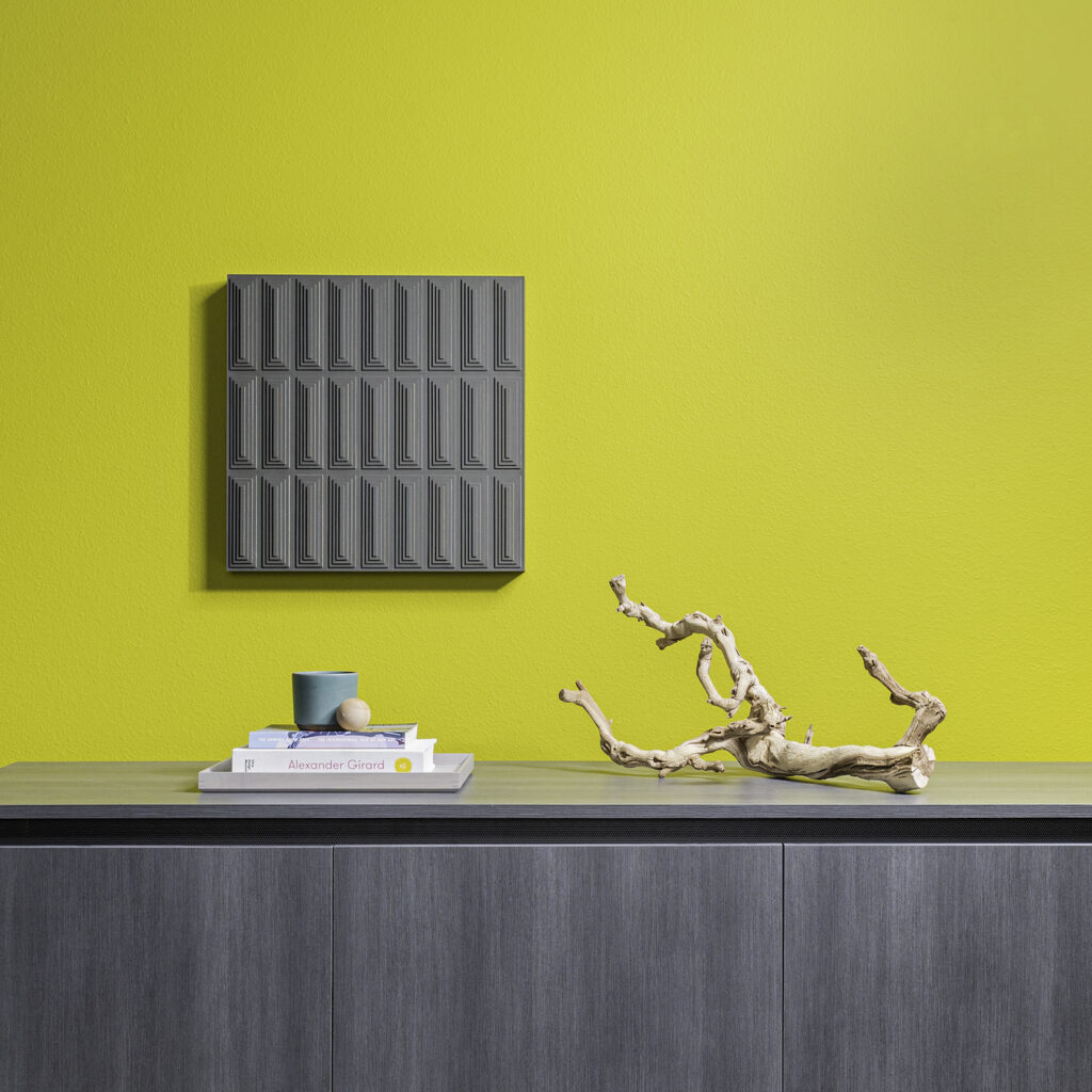 A square wall panel in gray leather. The surface is concentric rectangles on top of each other. It hangs on a chartreuse wall. In front of the wall is a gray cabinet with a pile of books and nicknacks and gnarled branch.