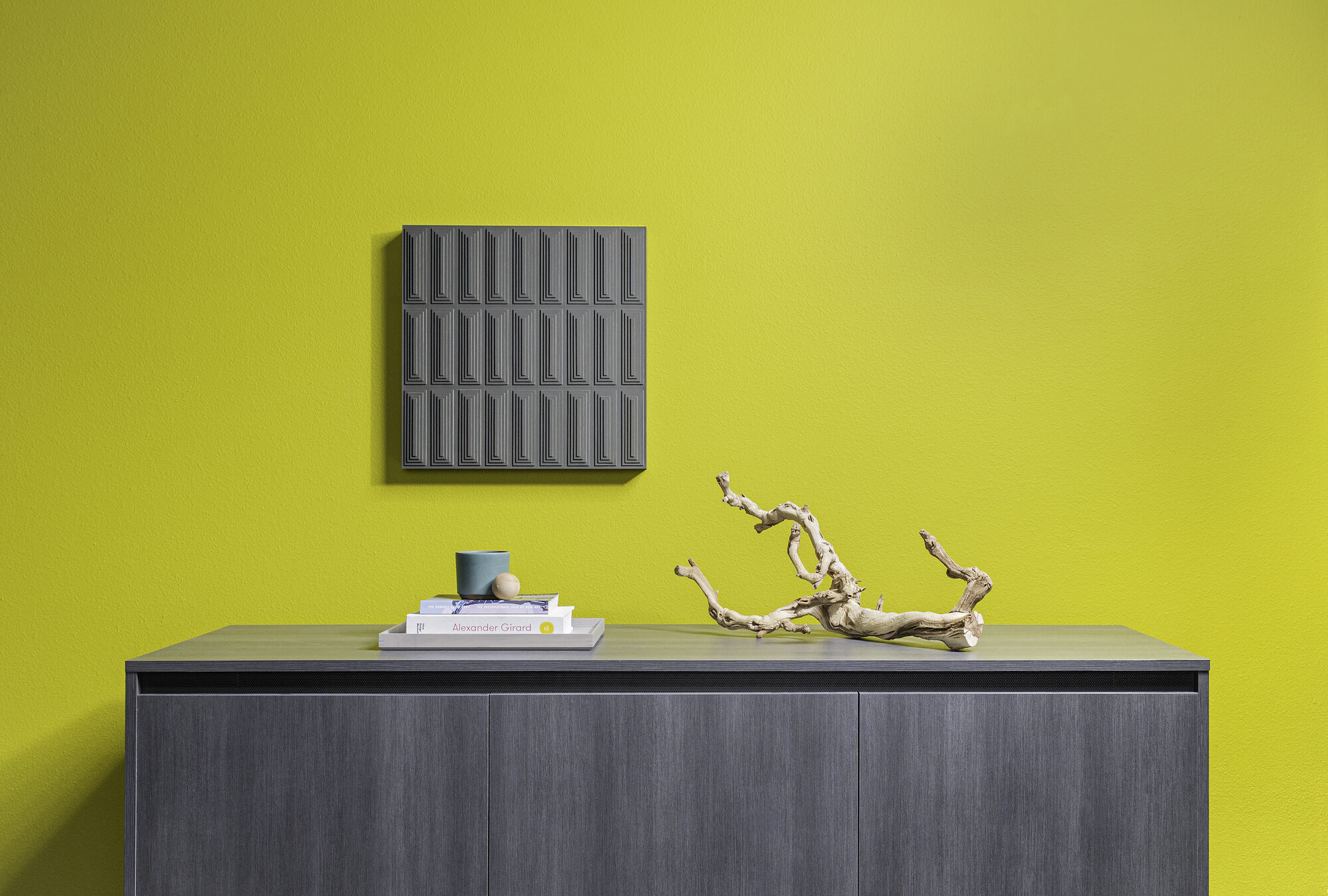 A long rectangular wall art piece hangs on a chartreuse wall. The piece has repeating diamond shapes in gray and the background felt is a bight orange.
