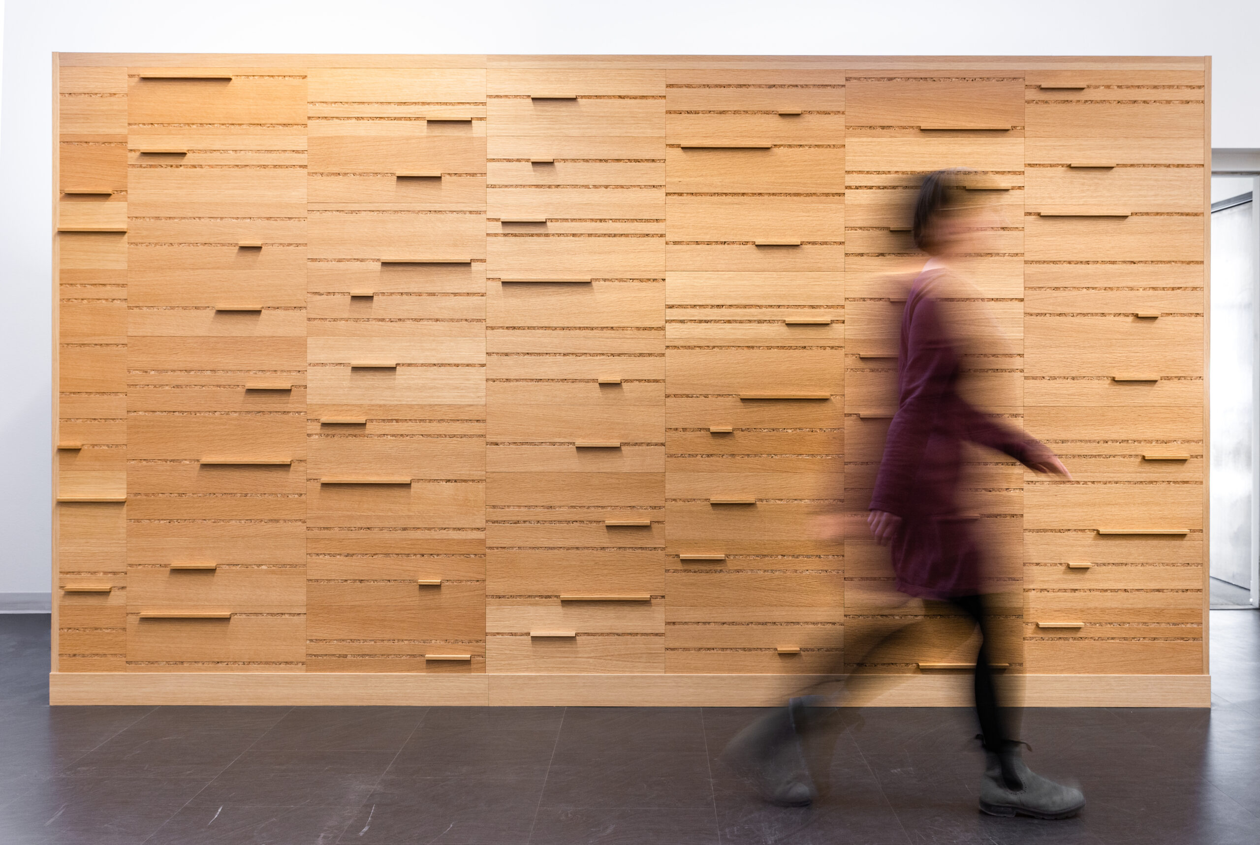 A blurry figure walks past an oak wood wall with horizontal cork lines and wooden bars set into the wall.