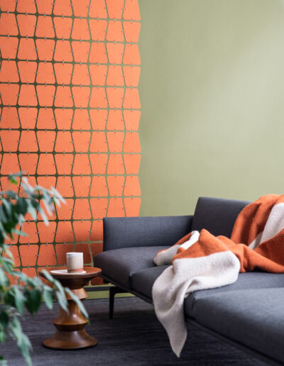 An orange room divider with soft trapezoidal shapes hangs to the left. There is a green wall, a gray couch with blanket, small table and mug and plant in front of the screen
