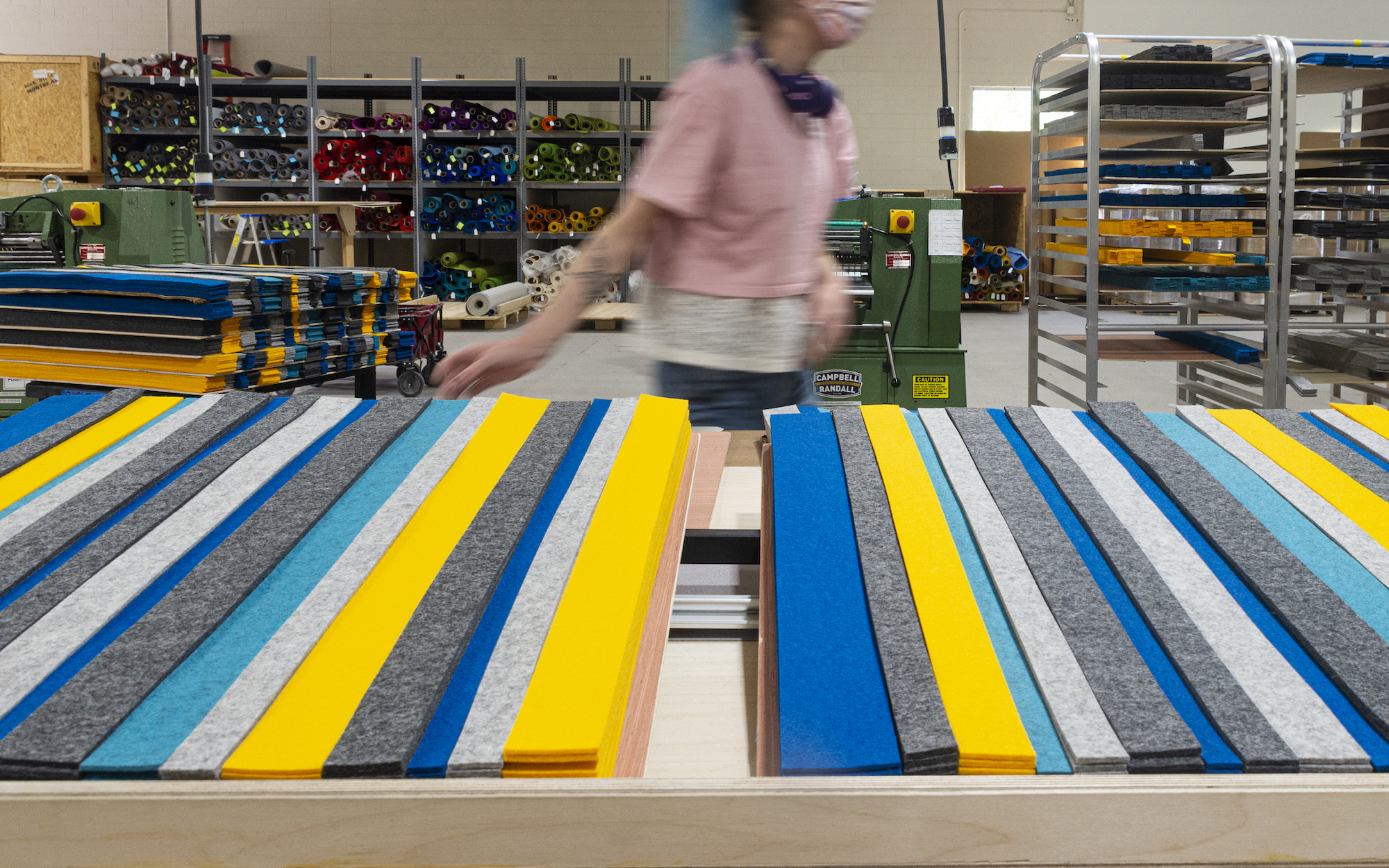 A person in a pink shirt walks past a table with blue, yellow, and gray strips of felt laid out. In the background are shelves with rolls of colorful felt