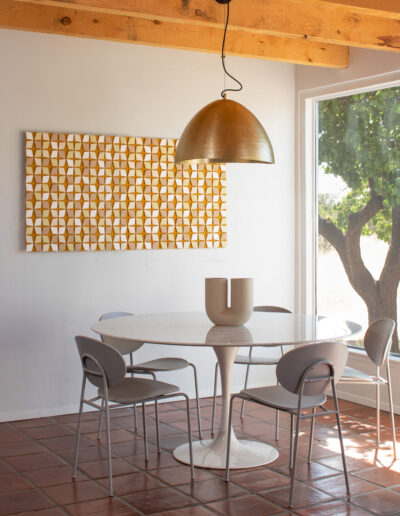 a felt wall art piece with diamond shapes in cream, light yellow and mustard colors, hangs in a dining space with a large window, round table, and hanging gold lamp