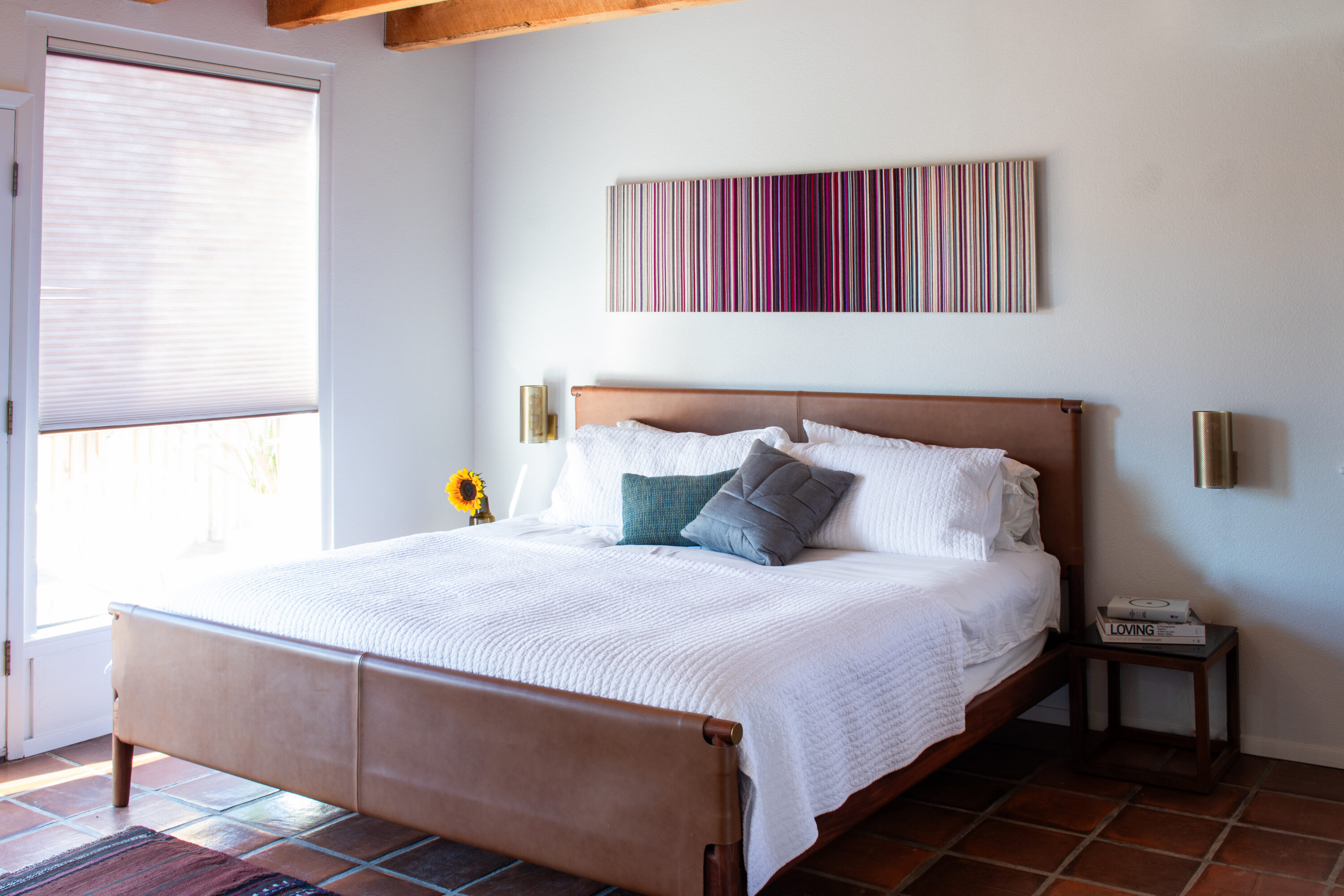a long rectangular felt panel with vertical strips of gradient color hangs above a bed with a blue blanket and two side tables.