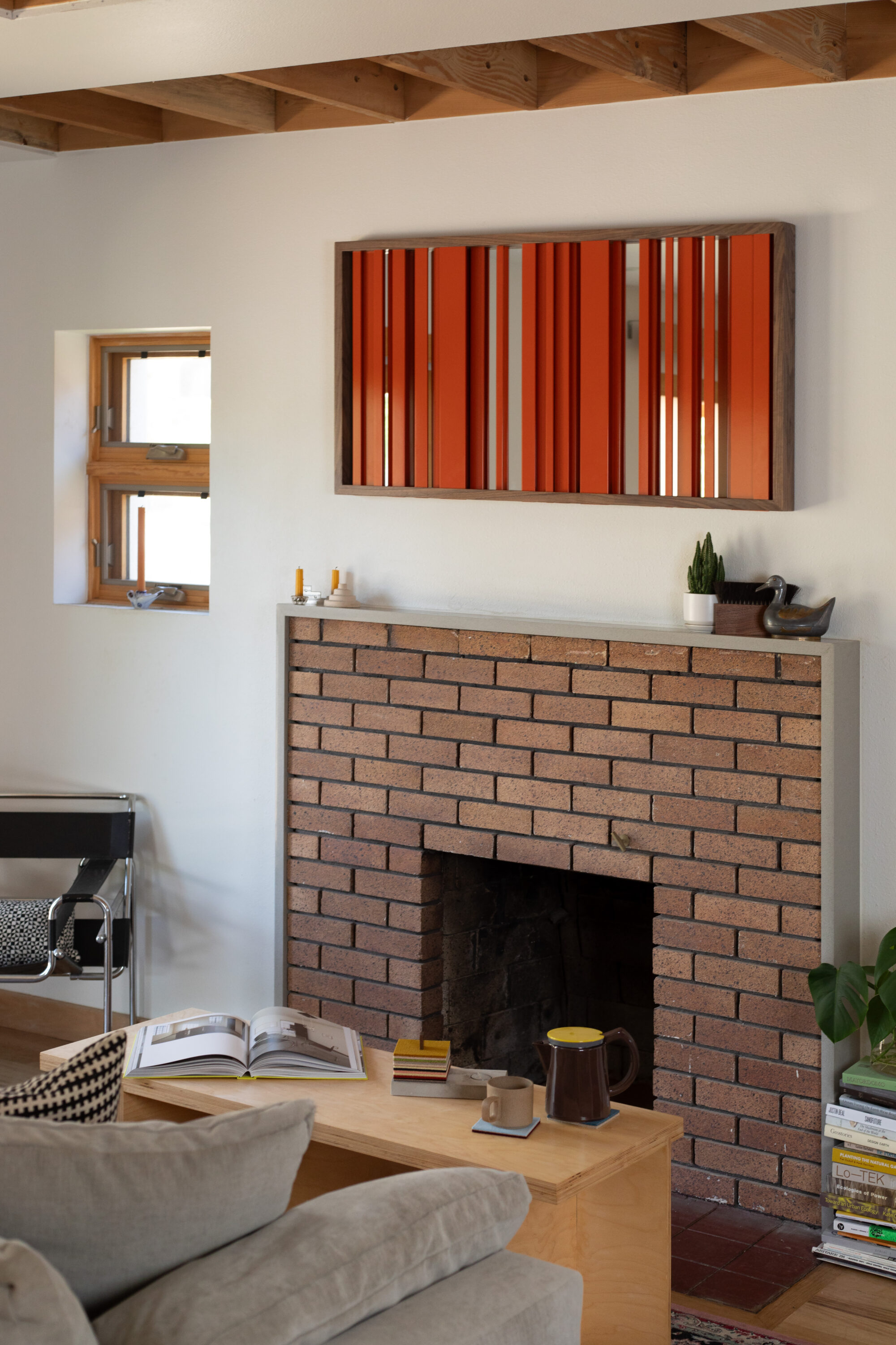 a living space in a modern home with a brick fireplace. above the fireplace hangs a horizontal rectangle. It is framed in dark wood, and red bands of metal are positioned vertically in the frame.