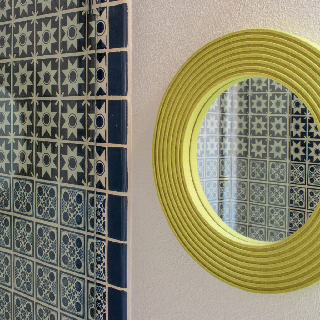 a round mirror framed with ribbed chartreuse wool felt hangs next to a tiled shower